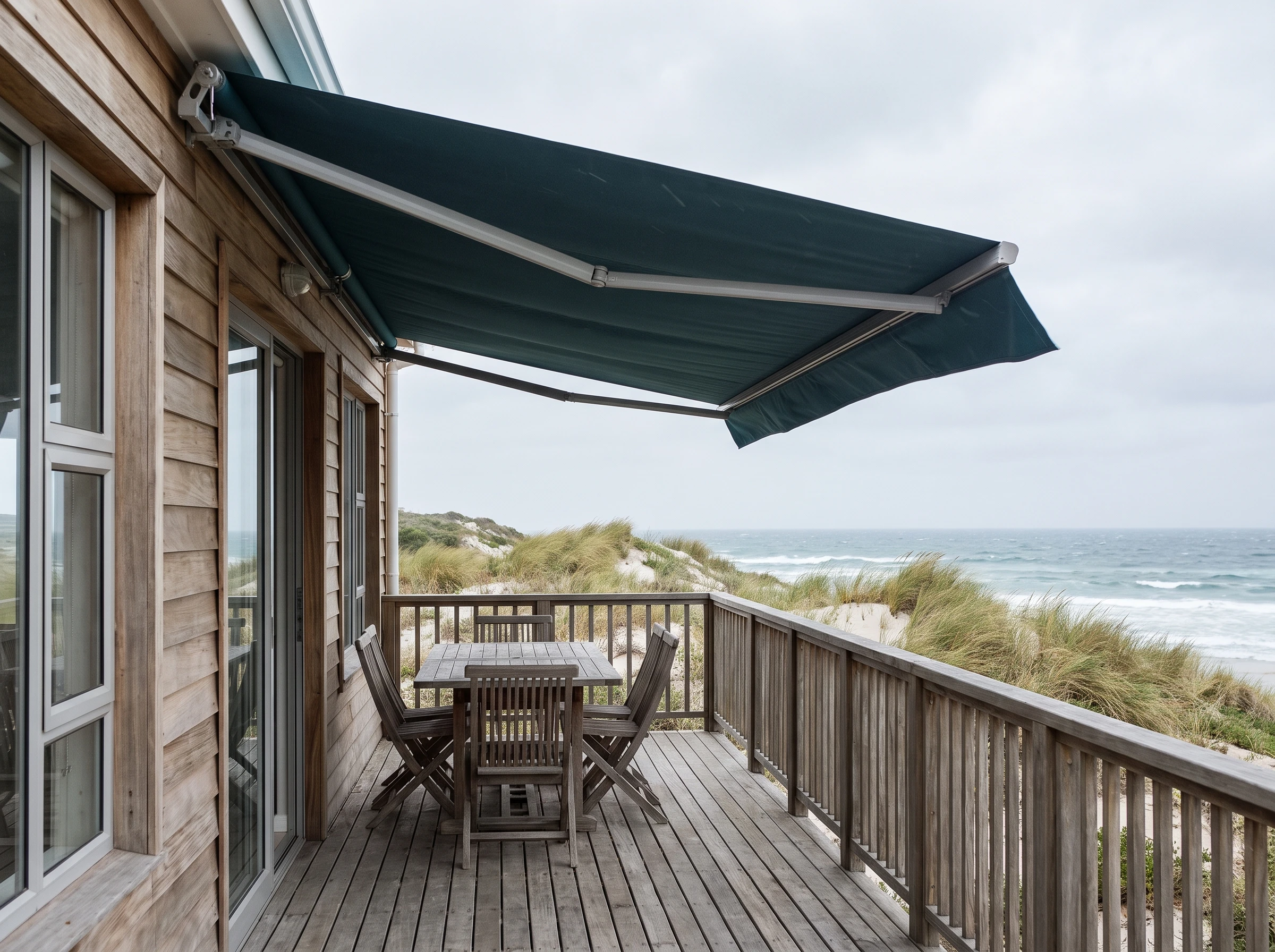 Retractable folding-arm awning in driftwood-tan fabric extended over an outdoor table on a Kommetjie beach house deck