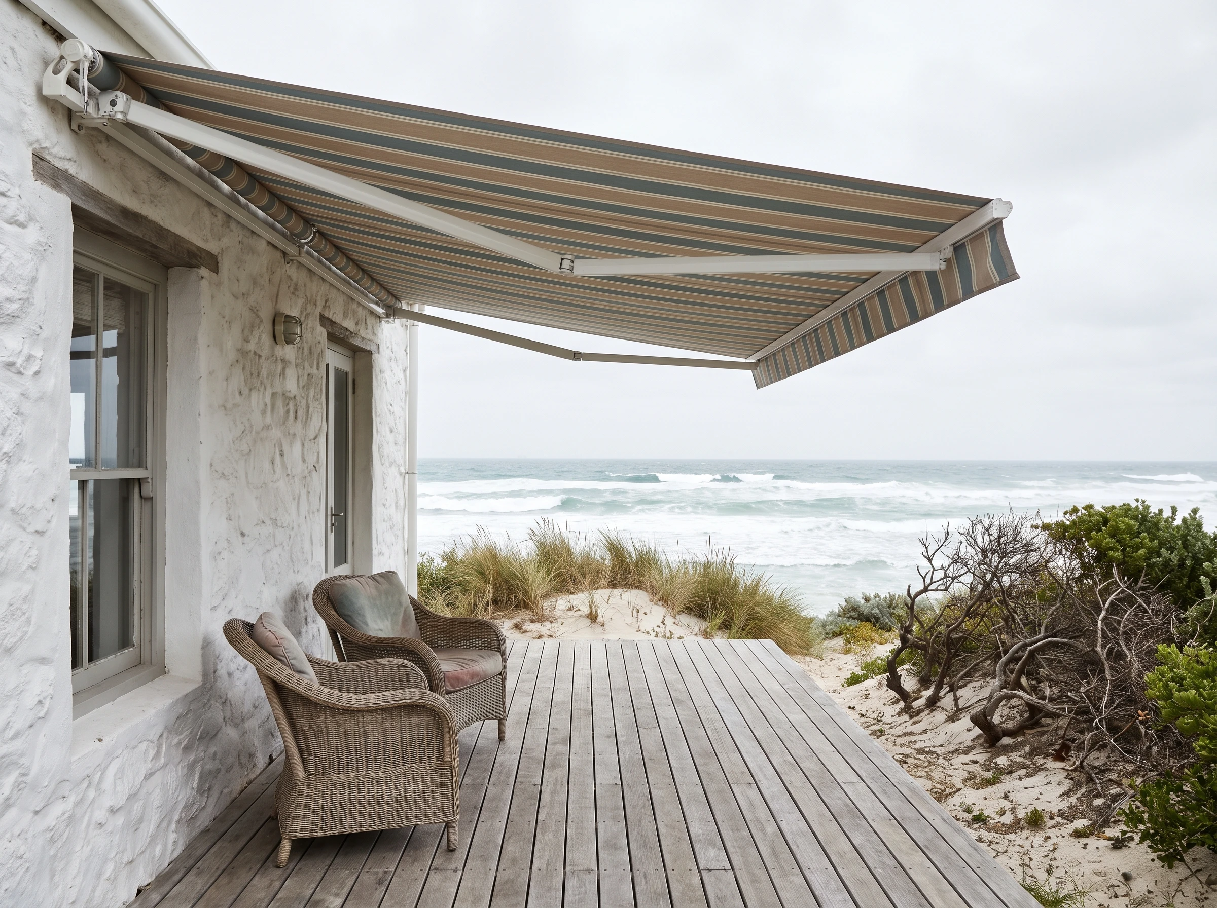 Folding-arm fabric awning fully extended over a wide timber stoep at a whitewashed Kommetjie cottage, wicker chairs beneath, dune and ocean beyond